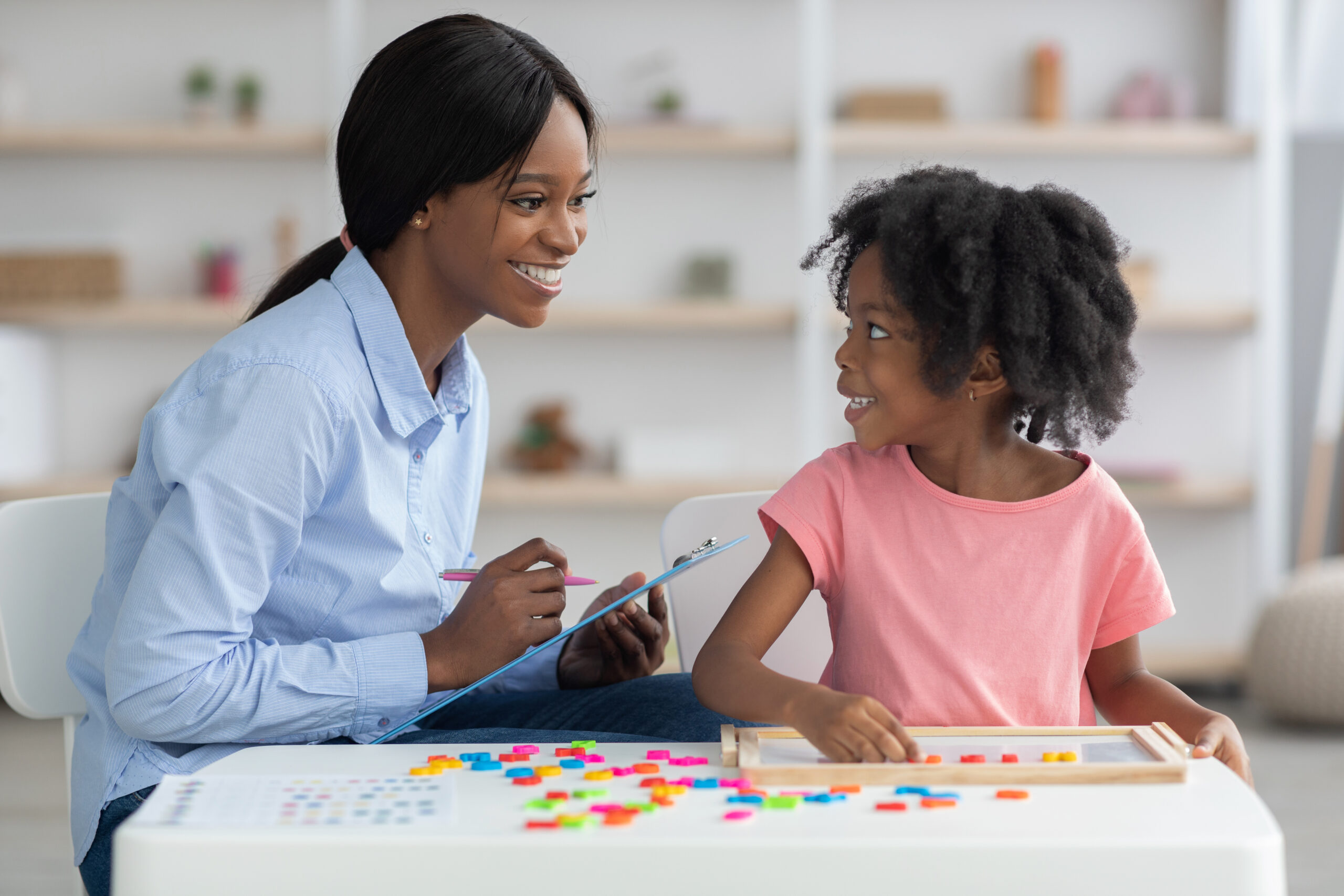 Cheerful child development specialist young african american lady testing cute little girl, sitting together at small table and playing with colorful plastic letters, black woman taking notes
