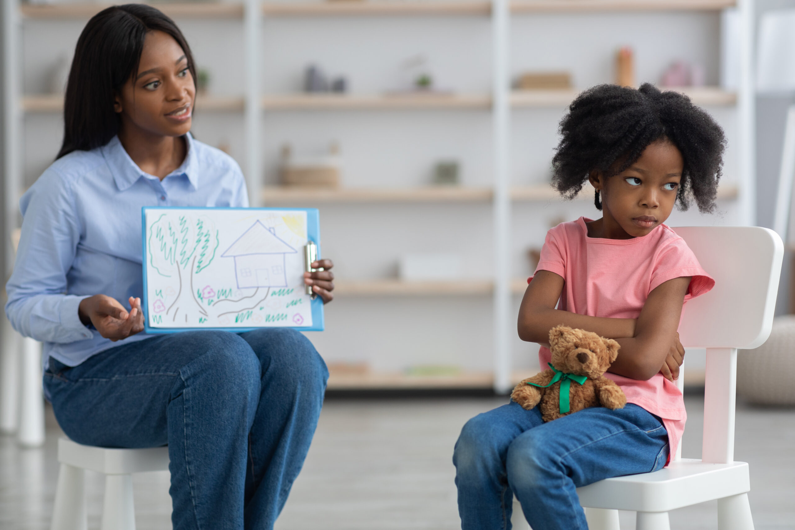 Defiant african american little girl with bushy hair and teddy bear avoiding young black woman child psychologist, kid looking aside while specialist showing drawing. Behavioral disorders in children